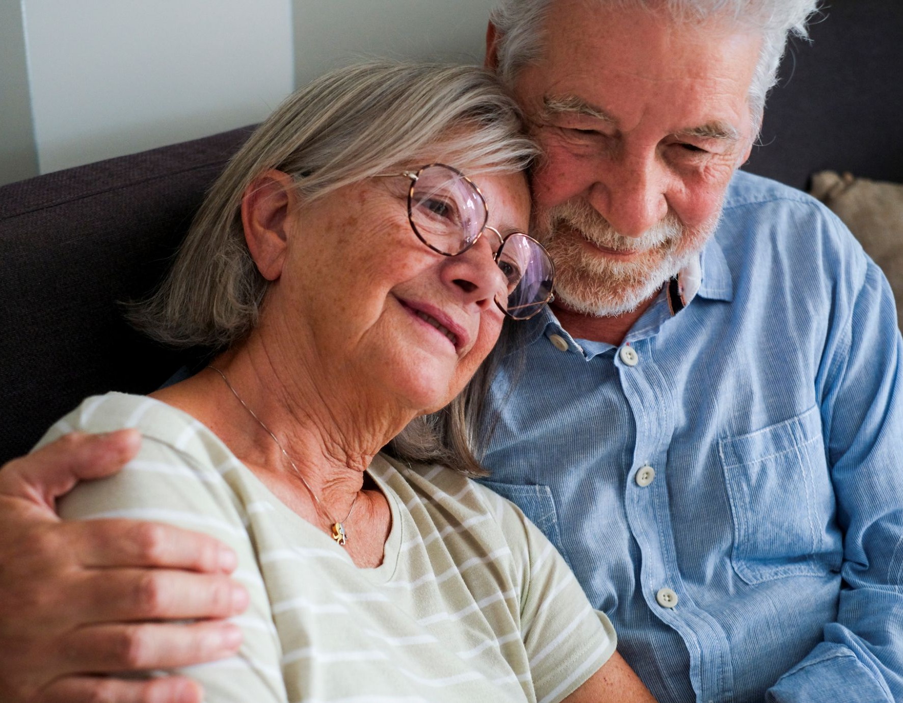 older adult couple sitting on a couch with his arm around her