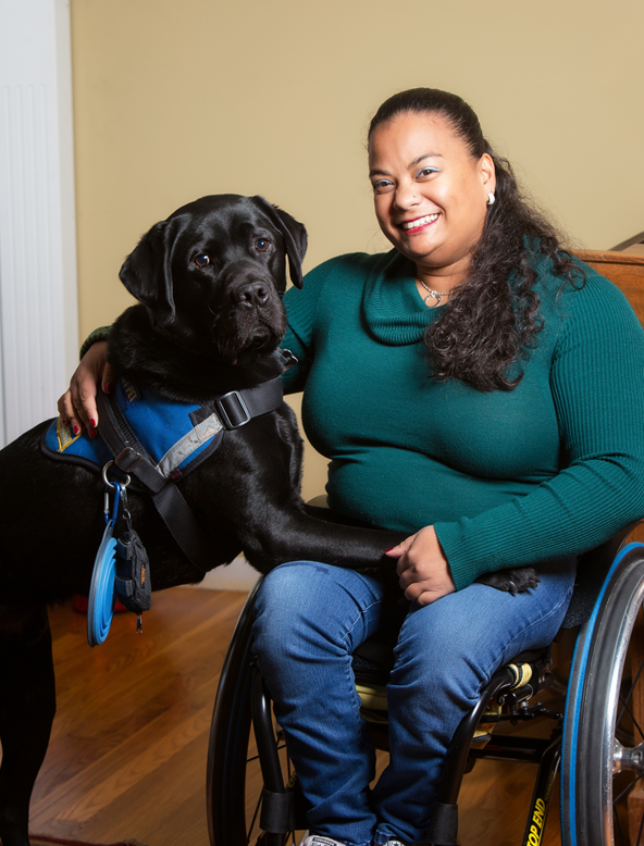 Photo of Anjali and her service dog, Kolton. Anjali has brown skin, brown eyes and long, curly black hair that she is wearing in a ponytail that drapes over her left shoulder. She is wearing blue jeans and a teal long-sleeved cowl-neck sweater. Kolton is a black lab wearing a blue and black harness. Anjali is sitting in her wheelchair, and Kolton is standing to her right with his front legs on her lap.