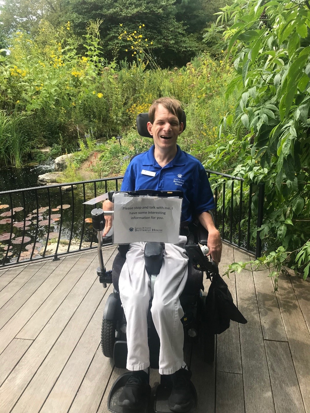 Jason with his wheelchair, AAC device and a sign inviting visitors to talk to him.