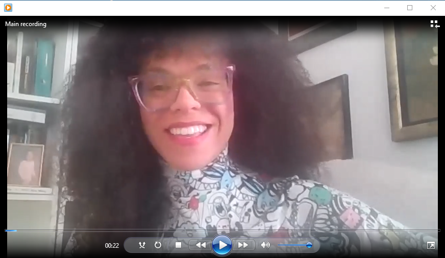  Reyma describes herself in this photo as a middle-aged, light-complected Black woman with shoulder-length, curly brown hair, wearing rainbow-colored glasses and a multi-colored turtleneck. She is seated in a white room with books and a picture of her daughter behind her. 