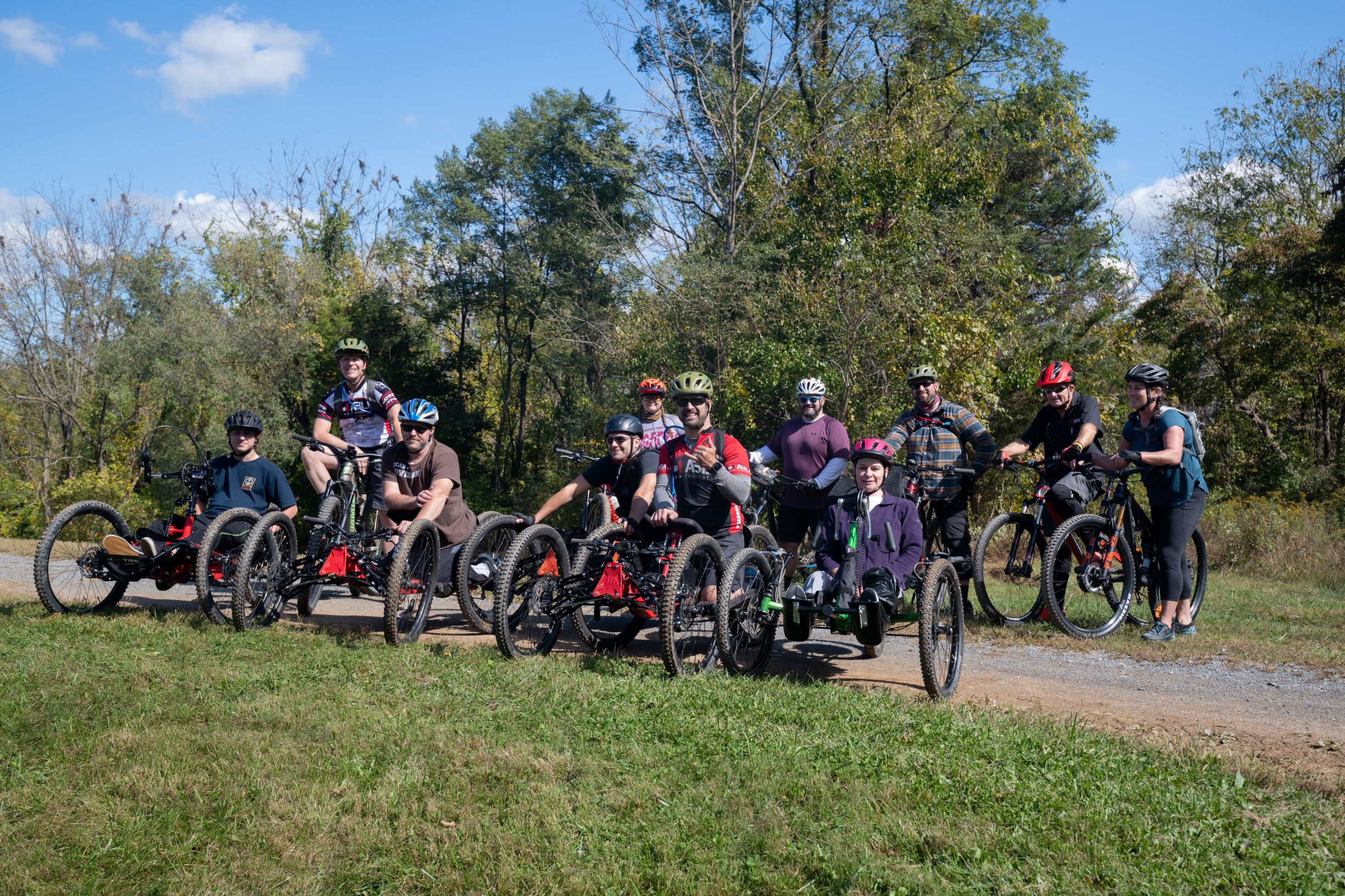 A group of smiling people on bicycles park on a field with the front row on adaptive bikes and a second row on regular bikes