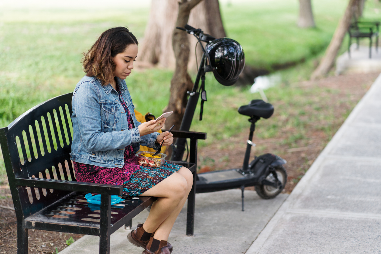 Woman eats her lunch on a bench, with a power scooter parked next to her